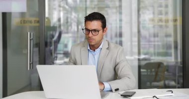 Odaklanmış genç İspanyol şirket CEO 'su dizüstü bilgisayarda yazıyor, ofis masasında çalışıyor, düşünceli oluyor, çeneye dokunuyor, başka yere bakıyor, iş planı düşünüyor, strateji yapıyor, online iletişim kuruyor.