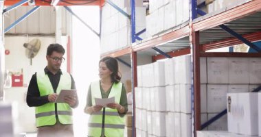 Erkek İspanyol depo müdürü kadın patrona rapor veriyor, metal raflarda yürüyor, kutuları işaret ediyor, depoyu gösteriyor. Stajyerleri erkek envanterine eğitmek, malları saymak, faturaları kontrol etmek.