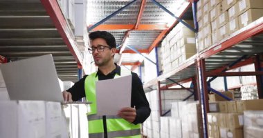 Hispanik depo çalışanı kağıt dağıtım faturalarını kontrol ediyor, depolama alanında dizüstü bilgisayar kullanıyor, teslimattan sonra kısmi numaralar giriyor, daktilo ediyor, danışmanlık kağıdı raporlarına bakıyor.