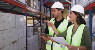 İki Hispanik depo çalışanı malları kontrol ediyor, ticareti tartışıyor, depodaki raflarda istiflenmiş kutuların başında duruyor, konuşuyor, faturaları ve tabletleri tutuyor.