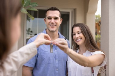 Yeni evlerinden anahtar alan, dışarıda emlakçıyla duran gülümseyen beyaz bir evli çift. Başarılı bir emlak anlaşması, ilk mülkün satın alınması, yazlık evde hafta sonu konaklama.