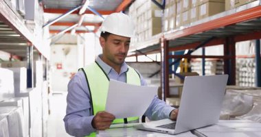 Diz üstü bilgisayarında çalışan, yol faturalarını kontrol eden, elektronik sevkiyata başvuran, depolama alanındaki bilgisayardaki lojistik uygulamayı kullanan, kasklı İspanyol erkek depo müdürü.