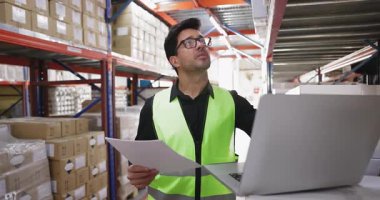 Ciddi bir Latin erkek depolama denetçisi teslimattan sonra malları sayıyor, envanter çıkarıyor, metal raflarda dizüstü bilgisayar kullanıyor, kutulara bakıyor, kağıt faturalarına ve bilgisayarlara bakıyor.