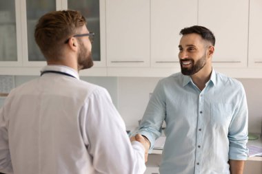 Tıbbi yardım için teşekkür ederim. Mutlu gülümseyen genç hasta güvenilir bir doktorla klinik muayene odasında tokalaşıyor. Profesyonel bakımdan sonra iyileştiği için şükrediyor.