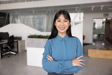 Mutlu, kendinden emin, 20 'li yaşlarda Asyalı genç bir iş kadını el ele tutuşup kameraya bakıyor, gülümsüyor, şirket ofisinde başarılı bir vesikalık fotoğraf çekimi için poz veriyor.