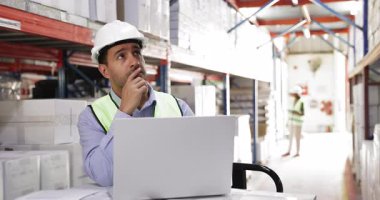 Ciddi genç Hispanik depo çalışanı, sert şapkalı, envanter yapıyor, metal raflardaki kutuları teker teker sayıyor, kağıt dağıtım faturalarına ve dizüstü bilgisayarlara danışıyor, endüstriyel depolamada çalışıyor.