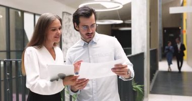 İki iş arkadaşı basılı proje belgelerini ve dijital tabletle ilgili verileri inceliyor, anahtar detayları tartışıyor, bilgiyi analiz ediyor, kararlar alıyor, şirket ofis koridorunda duruyor. Takım çalışması.