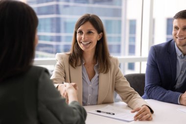 Orta yaşlı iş kadını toplantı masasında oturuyor, kadın müşteriyle tokalaşıyor, güven ve tatmin yayıyor, pazarlığı başarıyla sonuçlandırıyor, kurumsal ofiste iş anlaşması yapıyor.