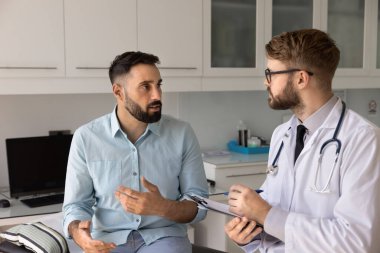 Güvenilir doktor randevusunda. Sağlık kontrolü için hastaneye giden genç bir erkek hasta muayene odasındaki koltuğa oturup GP ile ilgili endişelerini paylaşır muayene sırasında sağlık sorunlarını açıklar.