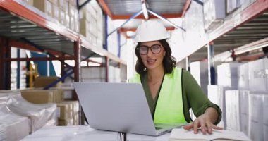 Depoda dizüstü bilgisayarda çalışan gözlüklü ve şapkalı genç, pozitif bir İspanyol kadın yönetici. Metal raflarda yığılmış kutularla birlikte bilgisayar kullanıyor. Defterde danışmanlık notları var.