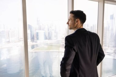 Olumlu düşünceli genç emlak yatırımcısı ofis penceresinden şehir görüşüne bakıyor, gayrimenkul projesini düşünüyor, yatırım planlıyor, iş molası veriyor, tatil yapıyor, hayal kuruyor.