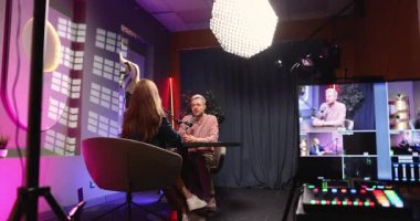 İki podcaster görüşme sırasında mikrofonla masada oturuyor. Stüdyo ışıkları, kameralar, kontrol monitörü canlı kayıt gösteriyor. Yaratıcı üretim, medya içeriği oluşturma, iletişim