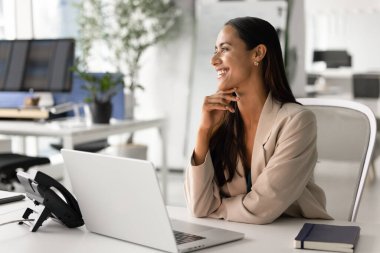 Zarif ceketli iyimser kadın çalışan, dizüstü bilgisayarla masada oturuyor, mutlu bir gülümsemeyle mesafeye bakıyor, kariyer fırsatı ve perspektifleri hayal ediyor. Gelecek, iş vizyonu, özlemler