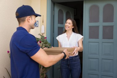 Kadın verandada duruyor, karton paket kutusunu alıyor. Yanında da erkek kuryeden gelen siparişler var. Gülümsüyor, hızlı teslimat hizmetlerinden ve başarılı online alışveriş deneyimlerinden memnun.