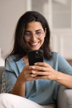 Gözlüklü güzel bir Latin kadın evdeki mobil uygulamalar aracılığıyla sohbet ediyor, akıllı telefonlu koltukta oturuyor, arıyor, web sörfü yapıyor, pazar platformunu kullanarak ürünler ve modern kıyafetler alıyor.