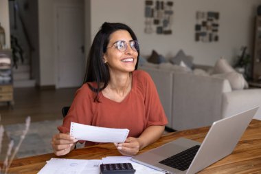 Gülümseyen Brezilyalı kadın faturasını tutuyor, evinde dizüstü bilgisayarla çalışıyor, kişisel mali işlerini başarıyla yürüttükten ve aile bütçesini dengeledikten sonra tatmin oluyor. Tasarruf, mali kilometre taşı elde etmek