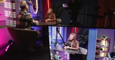 Profesyonel podcast kayıt oturumunun perde arkasında, masada sohbet eden iki sunucu, kameralar farklı açılar yakalar, monitör canlı medya prodüksiyonunu kapatır