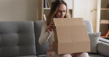 Kadın teslimat kutusunu açıyor, içine bakıyor, evde kendini üzgün hissediyor. Ürün tatminsizliği, geri dönüş isteği, geri ödeme, kalitesiz ürün, ekomünans sorunu, müşteri şikayeti, olumsuz alışveriş deneyimi