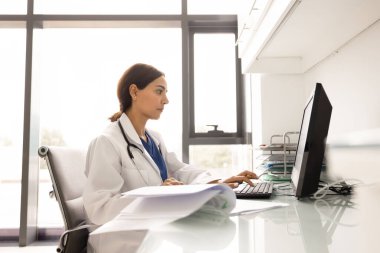 Modern klinik masasındaki masaüstü bilgisayarında çalışan beyaz önlüklü kadın doktor. Etrafı dokümanlar, tarih dergileri ve anestezilerle çevrili. Hasta kayıt yönetimi, tıbbi araştırma, sağlık hizmetleri
