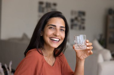 Güzel Brezilyalı mutlu bir kadının portresi, sıcacık bir iç mekanda elinde su bardağıyla poz veriyor, sağlıklı yaşam tarzını ve iyi yaşam alışkanlıklarını destekliyor, iyi sağlık ve güzellik için doğal su içiyor.