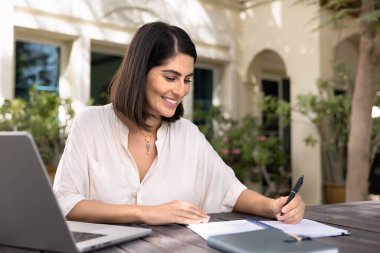 Laptopta çalışan, notlar yazan, evin yakınındaki bahçede oturan, gülümseyen, açık havada uzaktan çalışmaktan keyif alan, gülümseyen,