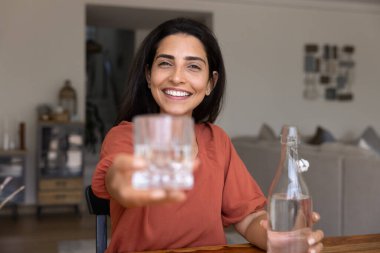 30 'lu yaşlarda genç bir Latin kadın kameraya bakıyor, kolunu uzatıyor, neşeli gülüşlü bir bardak doğal su sunuyor. Günlük su tüketiminin, susuzluğun, sağlığın ve sağlıklı alışkanlıkların önemi seçenekleri
