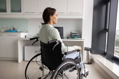 Tekerlekli sandalyedeki genç bir kadının muayene odasında oturması, geniş bir klinik penceresinden dikkatlice bakması, kazadan sonra rehabilitasyonun devamını düşünmesi, iyileşme umudu.