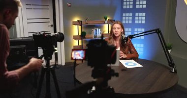 Gülümseyen kadın podcast sunucusu dizüstü bilgisayar ve mikrofonla masaya oturur, erkek kameraman kamerayı ayarlarken güvenli bir şekilde konuşur. Profesyonel yayın etkinliği, dijital medya üretimi iş başında