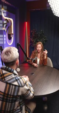 İki podcast sunucusu profesyonel mikrofonlar ve yaratıcı ışıklandırmayla donatılmış modern stüdyoda canlı yayında tartışıyorlar. Kadın konuşmacı ne demek istediğini açıklıyor, konuklarla sohbet ediyor. Yayın