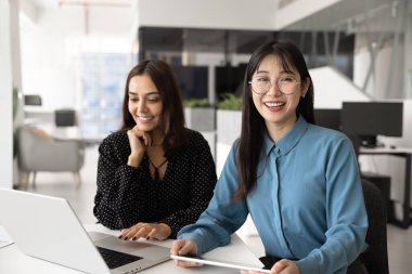 Takım çalışması için toplantı masasında poz veren neşeli, genç Asyalı proje yöneticisi kadın, kameraya bakıyor, gülümsüyor, Orta Doğulu meslektaşının yanında oturuyor dizüstü bilgisayarla yazı yazıyor.