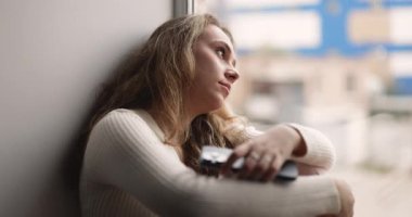 Üzgün, genç, yetişkin bir kadın mesaj ya da çağrı bekliyor. Pencere kenarında oturuyor, düşünceli bir şekilde pencereden dışarı bakıyor. Sıkılmış ya da yalnız hissediyor, hafta sonunu evde yalnız geçiriyor.