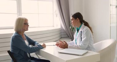 Beyaz önlüklü kadın doktor klinikteki masada oturur, sağlık tavsiyeleri verir, tedavi seçeneklerini açıklar, kadın hastalarla el sıkışır, tıbbi muayeneyi bitirir. Sağlık, güven.