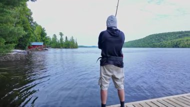 Tekne rıhtımından sakin gölde balık tutan adam olta atıyor ve makarayı kullanıyor. Spor balıkçısı UV koruyucu başlık ya da kar maskesi olarak kar maskesi kullanıyor. Balık tutma yaz tatili