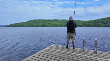 Tekne rıhtımından sakin gölde balık tutan adam olta atıyor ve makarayı kullanıyor. Spor balıkçısı UV koruyucu başlık ya da kar maskesi olarak kar maskesi kullanıyor. Balık tutma yaz tatili