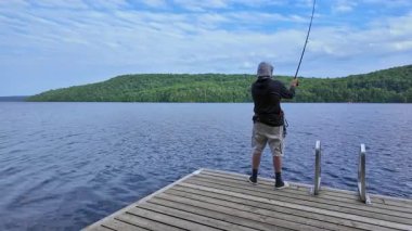 Tekne rıhtımından sakin gölde balık tutan adam olta atıyor ve makarayı kullanıyor. Spor balıkçısı UV koruyucu başlık ya da kar maskesi olarak kar maskesi kullanıyor. Balık tutma yaz tatili