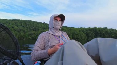 Huzurlu gölde balık tutmak için PDL kayağında pedal çeviren bir adam. Balıkçılık sporcusu UV koruyucu başlık ya da kar maskesi olarak kar maskesi kullanıyor. Balık tutma yaz tatili.