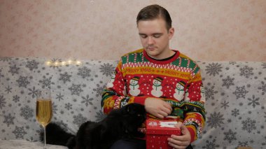 Yeni yıl tatili genç adam bir hediye açar ve paketler, şampanya, yeni yıl hediyeleri, yeni yıl süsleri ve iyi bir ruh hali ve siyah bir kedi.