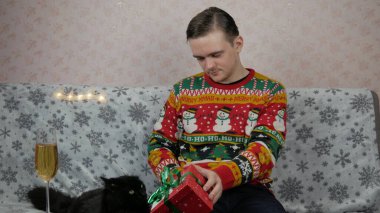 Yeni yıl tatili genç adam bir hediye açar ve paketler, şampanya, yeni yıl hediyeleri, yeni yıl süsleri ve iyi bir ruh hali ve siyah bir kedi.