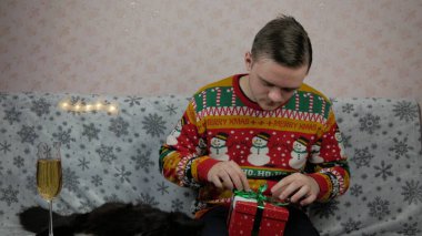 Yeni yıl tatili genç adam bir hediye açar ve paketler, şampanya, yeni yıl hediyeleri, yeni yıl süsleri ve iyi bir ruh hali ve siyah bir kedi.