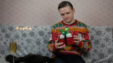 Yeni yıl tatili genç adam bir hediye açar ve paketler, şampanya, yeni yıl hediyeleri, yeni yıl süsleri ve iyi bir ruh hali ve siyah bir kedi.