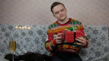 Yeni yıl tatili genç adam bir hediye açar ve paketler, şampanya, yeni yıl hediyeleri, yeni yıl süsleri ve iyi bir ruh hali ve siyah bir kedi.