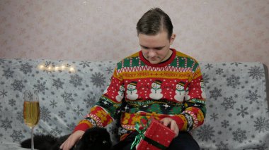 Yeni yıl tatili genç adam bir hediye açar ve paketler, şampanya, yeni yıl hediyeleri, yeni yıl süsleri ve iyi bir ruh hali ve siyah bir kedi.