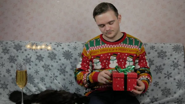 Yeni yıl tatili genç adam bir hediye açar ve paketler, şampanya, yeni yıl hediyeleri, yeni yıl süsleri ve iyi bir ruh hali ve siyah bir kedi.