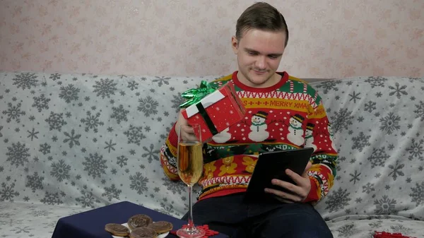 Yeni yıl tatili, genç bir adam bir hediye açar ve paketler, şampanya, yeni yıl hediyeleri, yeni yıl süslemeleri ve aile ile bir sohbet, online tablet