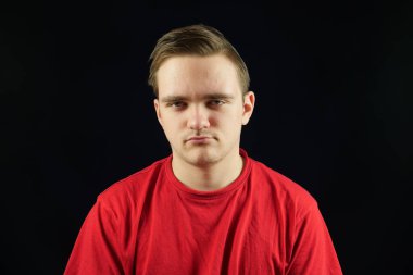 Sıradan tişörtlü genç yakışıklı, soyutlanmış karanlık arka planda duruyor, kızgın ve düşünceli, somurtkan yüzlü.