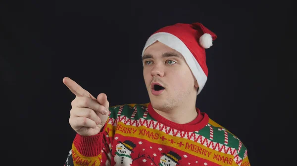 Genç yakışıklı adam duygularını gösterir - parmağını gösterir, kırmızı bir yeni yıl süveteri giyer ve yeni yıl çizimleri ve kırmızı bir şapka giyer.