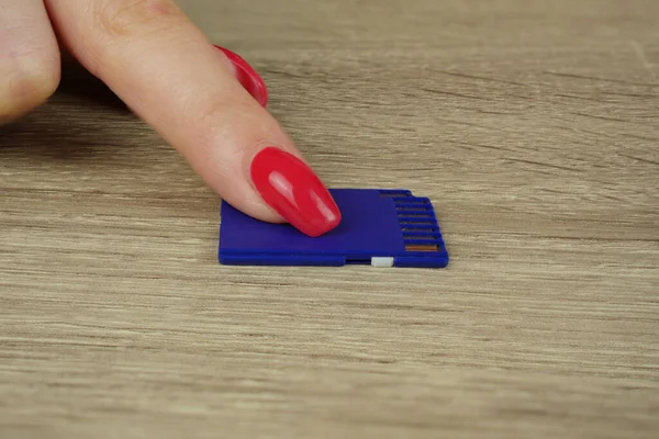 Kamera için mavi bir hafıza kartı masanın üzerinde duruyor ve kırmızı manikürlü bir kadının parmağı parmağıyla karta basıyor.