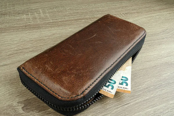 Üzerinde euro banknotları olan kahverengi deri bir cüzdan masanın üzerinde duruyor ve banknotlar cüzdanın içinde görünüyor.