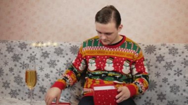 Yeni yıl süveteri giyen bir adam kanepeye oturur ve kırmızı bir kutuya hediye koyar, oyuncak ve tıbbi bir maske takar, bir bardak şampanyanın yanında, hediyeyi hangi kurdeleyle paketleyeceğini seçer, kırmızı mı yeşil mi?