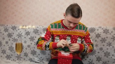 Yeni yıl süveteri giyen bir adam kanepeye oturur ve kırmızı bir kutuya hediye koyar, oyuncak ve tıbbi bir maske takar, bir bardak şampanyanın yanında, hediyeyi hangi kurdeleyle paketleyeceğini seçer, kırmızı mı yeşil mi?